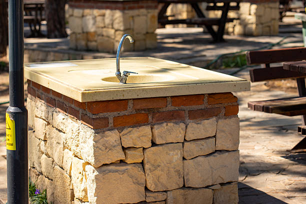 A stone water fountain stands in a park. Picnic tables are visible in the background, with sunlight dappling the scene.