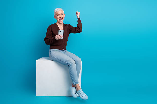 istock Happy senior woman using smartphone while sitting on a studio block with a bright blue background showcasing excitement 2236041873