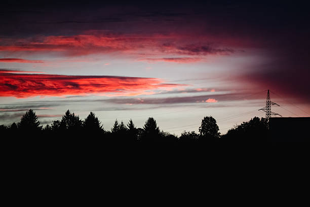 Vivid red and purple clouds stretch across the evening sky above a dark forest silhouette, with a single power line tower on the right. The striking contrast between nature and infrastructure evokes a dramatic and atmospheric twilight scene.