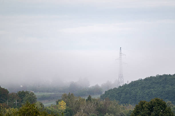 A misty rural landscape featuring a tall transmission tower rising above the fog. The soft haze partially covers the trees and hills, blending natural and industrial elements in a calm, atmospheric scene.