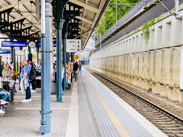 Gdańsk Główny - Empty train tracks run alongside the platform at Gdansk main train station, where passengers wait under a roofed section. People sit and stand near blue metal pillars, while the wall along the track creates a narrow view down the line. Electronic displays and signage indicate train schedules.