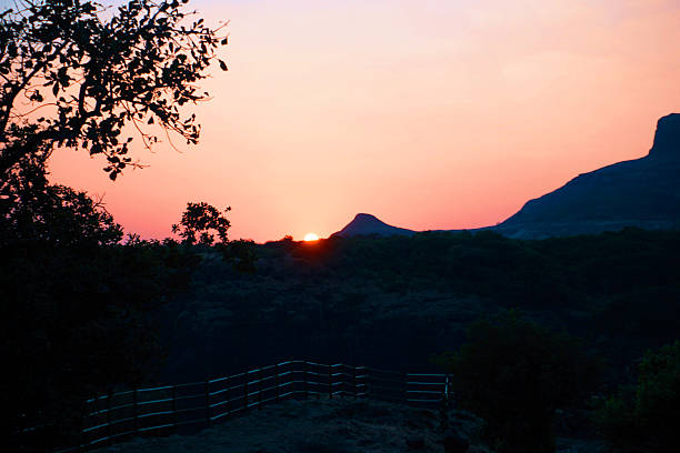 Tranquil landscape of the sun cresting a distant ridge, casting pink-orange light across the sky. A metal railing frames a scenic viewpoint in the foreground, with dark trees and rolling hills creating layered silhouettes. Ideal for themes of travel, hiking, national parks, dawn/dusk, serenity, and nature backgrounds.