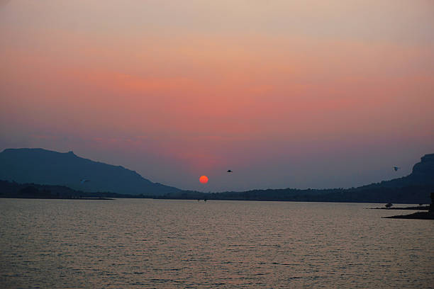 Tranquil scene of the sun setting over a calm lake, casting pastel pink and orange hues across the sky. Distant hills frame the horizon while a few birds glide above the water, creating a peaceful, contemplative mood—ideal for themes of nature, travel, relaxation, mindfulness, and evening landscapes.