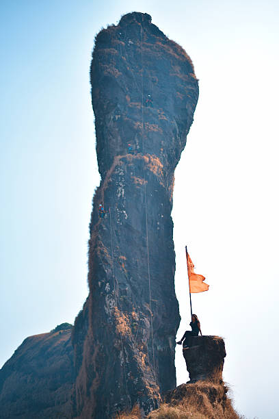A businessman stands at the top of a mountain peak as he holds a large orange flag attached to a pole. He stands as he looks out into the distance proudly proclaiming his achievement.
