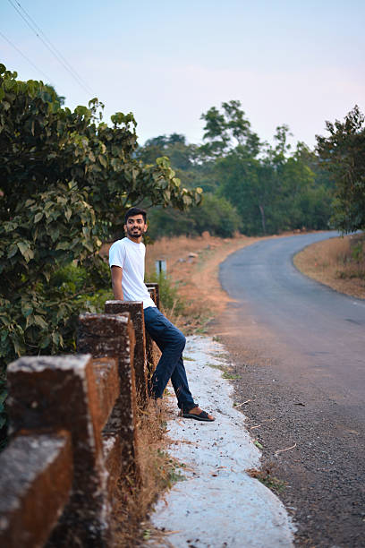 Outdoor portrait of a smiling young man perched on a weathered bridge railing next to a quiet rural road that curves into green trees at dusk. Relaxed travel vibe ideal for road trip, freedom, weekend getaway, and outdoor lifestyle themes.