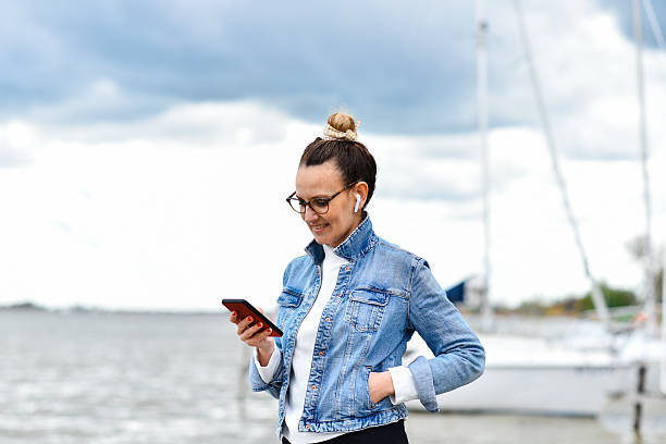istock Smiling Woman Using Smartphone Outdoors by the Water on a Cloudy Day 2236053081