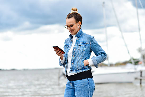 istock Smiling Woman Using Smartphone Outdoors by the Water on a Cloudy Day 2236053689