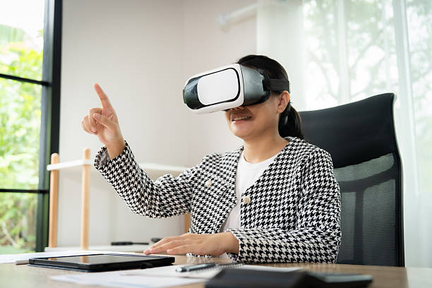 istock A woman wearing a black and white jacket is pointing at something while wearing a virtual reality headset. She is sitting at a desk with a laptop and a tablet 2236053769