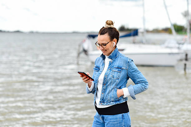 istock Smiling Woman Using Smartphone Outdoors by the Water on a Cloudy Day 2236053943