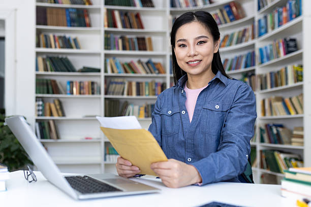 A smiling Asian woman holds documents in a library setting. Bookshelves filled with books create a backdrop.