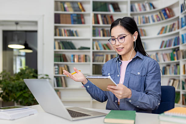 A young Asian woman participates in a video call while working remotely from a library. An Asian woman is on a video call in a library, gesturing with a pencil, taking notes, and using her laptop. online tutors stock pictures, royalty-free photos & images