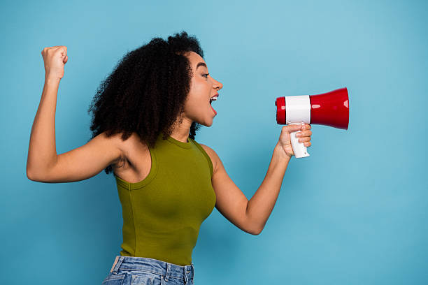 istock Young woman holding megaphone enthusiastically expressing message with gesture on blue background wearing trendy outfit 2236059842