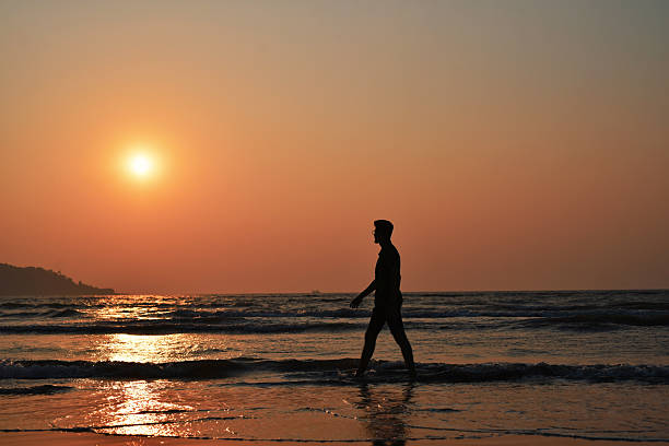 Peaceful coastal scene of a lone man strolling through ankle-deep surf as the sun sets over gentle waves. Warm orange sky and shimmering reflections on wet sand evoke themes of freedom, mindfulness, travel, coastal lifestyle, and golden-hour serenity.