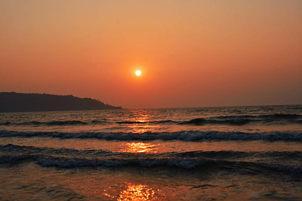 Warm orange sun sets above a calm sea, casting shimmering reflections across rolling waves with a distant headland in silhouette. Peaceful coastal seascape ideal for themes of travel, relaxation, mindfulness, nature backgrounds, summer vacation, and golden hour.