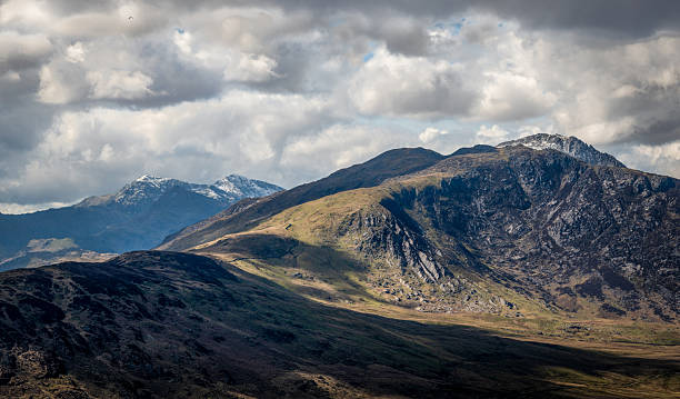 Rugged peaks rise under a sky of shifting clouds in the Snowdonia mountains of North Wales. Light and shadow sweep across the slopes, highlighting green ridges and rocky crags, while snow lingers on the higher summits in the distance. This dramatic scene captures the wild and untamed beauty of Eryri National Park, a landscape shaped by nature and time.