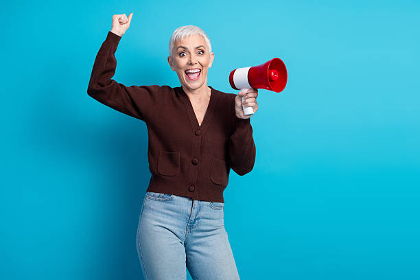 istock Happy elderly woman holding a megaphone against a bright blue background, expressing excitement and vibrant energy 2236078466