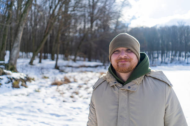 A man stands in a snowy forest. Trees surround him, and the winter air is crisp and cool. His warm clothing contrasts with the cold surroundings.
