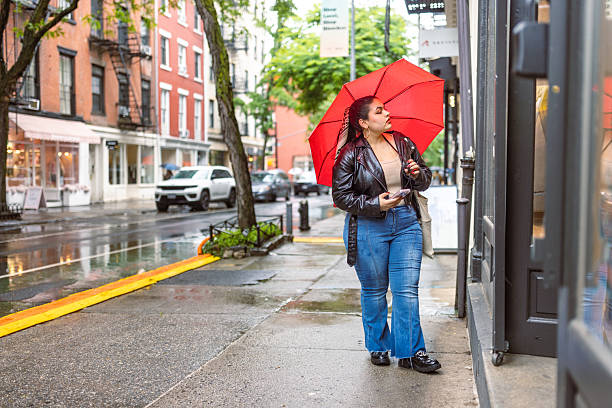 A woman wearing a black jacket and blue jeans walks on a rainy city street with a red umbrella, stopping to look into a shop window.