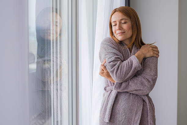 istock Serene woman wearing bathrobe hugging herself by the window 2236086515