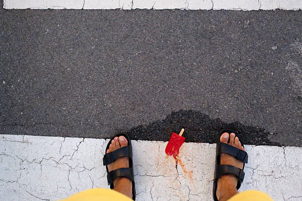 Point of view of a person wearing sandals and yellow shorts looking at a red popsicle melting on asphalt on a pedestrian crossing during a hot summer day