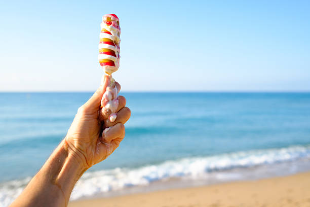 Hand holding a colorful ice cream on a stick, melting under the hot summer sun at the beach, with the sparkling sea and gentle waves creating a vibrant backdrop