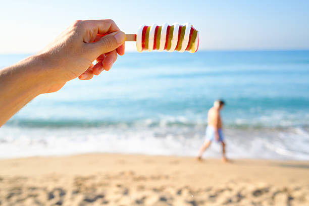 Colorful popsicle is melting quickly in the summer heat as someone holds it up against the backdrop of a beautiful beach with a blurred figure walking along the shore