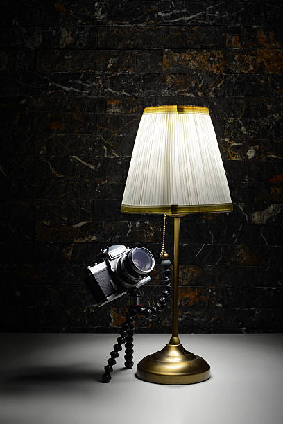 A creative surreal composition of a vintage film camera attached to a flexible tripod that resembles octopus-like legs, giving the impression of a living creature. The camera appears to reach out and pull the chain of a brass table lamp with a white lampshade, illuminating the dark scene. The playful setup makes the camera look alive, curiously gazing at the light it has just turned on. A whimsical and imaginative concept blending photography, humor, and surreal art.