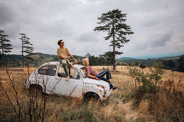 Couple sitting on the roof of an old rusty car, enjoying a peaceful moment in the countryside.
