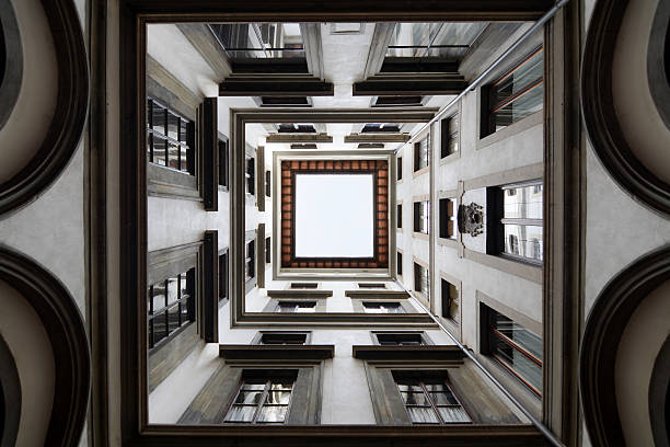 Looking upward from the inner courtyard of a historic building in Florence, Italy. The architectural symmetry of the walls, windows, and stone frames creates a striking square composition around the pale sky above.