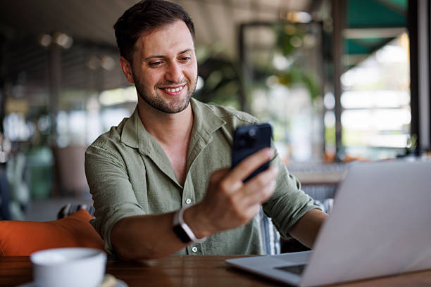 istock Young smiling man using mobile phone while working on a laptop at a cafe 2236096958