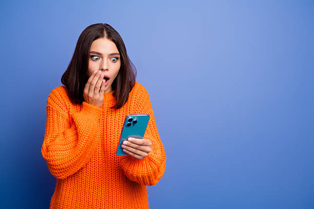 istock Surprised young woman in orange sweater using smartphone against blue background showing expressive emotion 2236100433