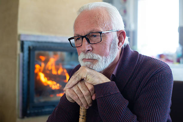 Senior man using walking cane sitting in his home in winter in front of the fireplace.