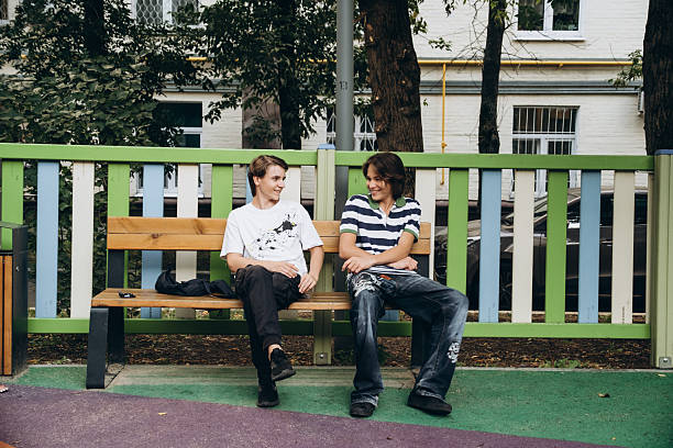 Two teenagers sitting on a park bench, engaged in cheerful conversation. Their expressions reflect joy and friendship, creating a warm and relaxed atmosphere in the outdoor setting.