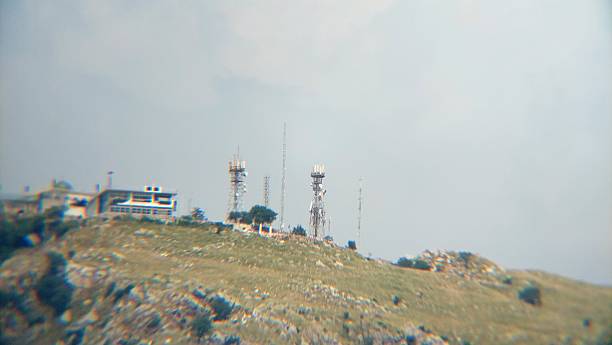 Hill summit with telecommunication towers and a building under a hazy, overcast sky. The landscape features a grassy, rocky terrain with scattered bushes. The towers are metal structures, equipped with antennas for communication purposes. The building, modern in design, is situated near the towers. The overall scene conveys a blend of technology and natural environment, with the cloudy sky adding a muted backdrop.