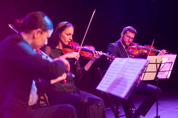 A diverse trio of professional violinists performs passionately on stage, illuminated by soft purple lighting, during an evening classical music concert.
