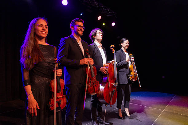 A diverse string quartet, including two men and two women, performing with violins and cellos on a brightly lit concert stage.