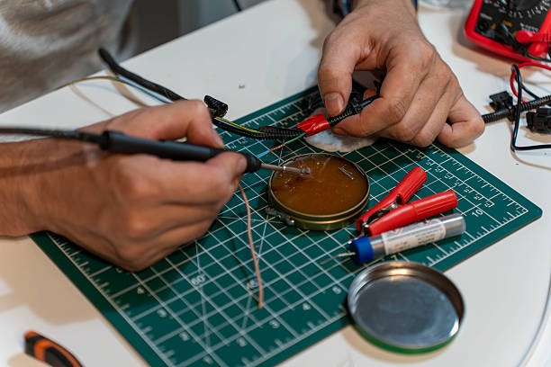 Detail view of human hands soldering electrical wires on a green cutting mat with soldering iron and repair tools. Concept of technical service and craftsmanship.