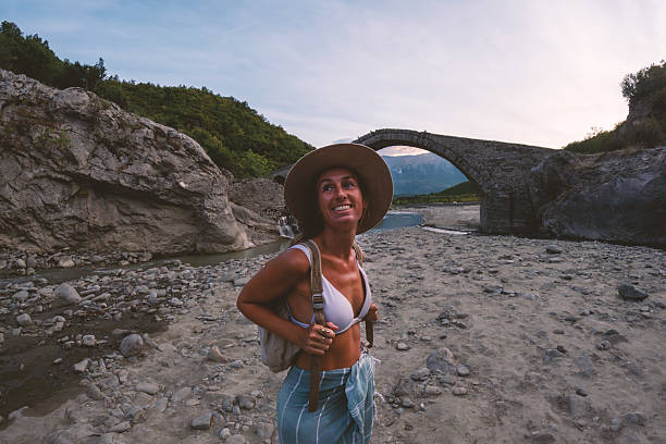 Smiling woman in hat and backpack hiking on rocky riverbed near historic stone arch bridge. Outdoor adventure traveler exploring nature and cultural landmarks. Active lifestyle and tourism.