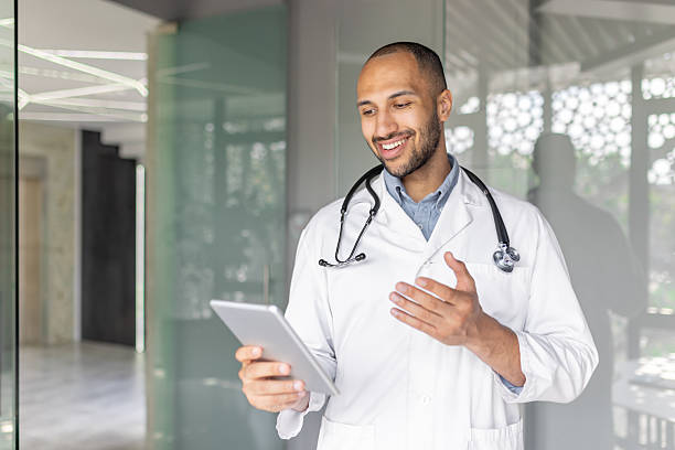 istock Young doctor consults patients remotely using tablet computer and video call. Man in white medical coat talking while standing inside office of modern clinic 2236117027
