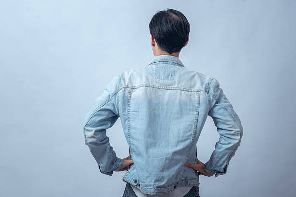 istock Asian man in denim jacket standing with hands on hips, seen from behind, posing in studio against plain light background, showing confident stance and casual modern fashion style 2236126661