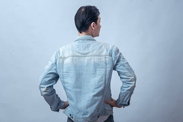 istock Asian man in denim jacket standing with hands on hips, seen from behind, posing in studio against plain light background, showing confident stance and casual modern fashion style 2236126663