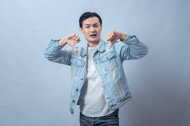 istock Asian man in denim jacket showing two thumbs down with a displeased expression, expressing dislike or disapproval in front of a plain studio background 2236126884