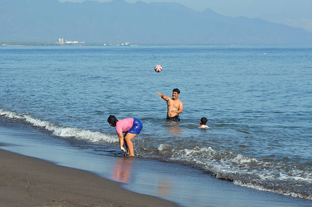 The father throws a ball high above as his son plays in shallow water and his daughter enjoys splashing at the foamy edge of the beach shoreline.