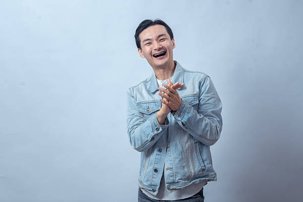 istock Asian man in denim jacket laughing with joy, clapping his hands together while smiling brightly in a casual studio portrait against a plain background 2236127315
