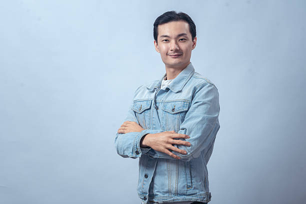 istock Asian man in light blue denim jacket standing with arms crossed against plain studio background, looking at camera with calm confident expression for lifestyle or business concept 2236127761
