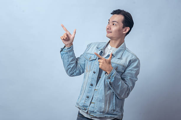 istock Asian man in denim jacket pointing upward and sideways with both hands against plain studio background, showing direction gesture with confident expression for lifestyle or business concept 2236127786