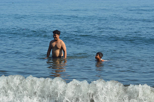 A father walks happily in shallow seawater, smiling at his son who floats with a ball, surrounded by calm waves and a peaceful shoreline.