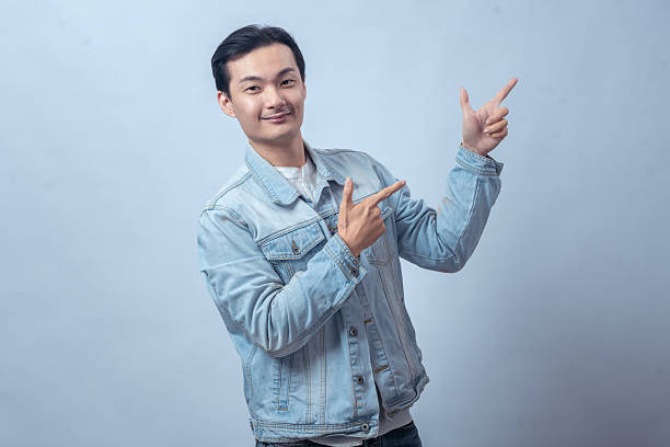istock Asian man in denim jacket pointing upward and sideways with both hands against plain studio background, showing direction gesture with confident expression for lifestyle or business concept 2236127807