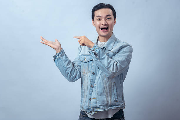 istock Asian man in denim jacket pointing upward and sideways with both hands against plain studio background, showing direction gesture with confident expression for lifestyle or business concept 2236127834