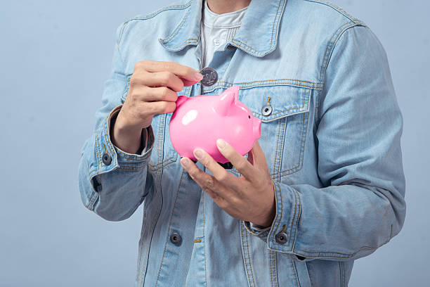 istock Asian man wearing a denim jacket smiling while inserting a coin into a pink piggy bank against a plain background, symbolizing saving money and financial planning 2236128907